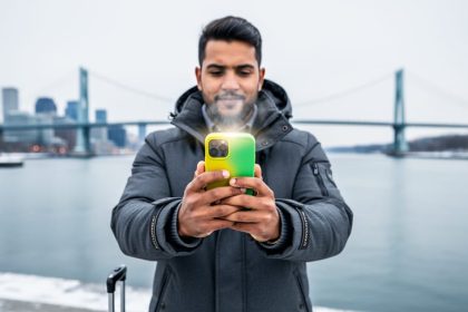Traveler in a winter coat holds a smartphone with a green-and-yellow case next to a carry-on at Montreal’s Old Port, with the city skyline and Jacques Cartier Bridge softly out of focus.